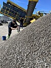 En arbeider står ved en stor haug med grått knust steinmateriale under et gult knuseanlegg.