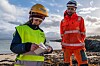 To personer i verneklær står ved sjøen; den ene holder en liten flaske ved en steinete strand.