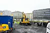 En CAT-gravemaskin står på en gruslagt byggeplass omgitt av bygninger og sperringer i et bysentrum.